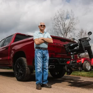 A man standing net to his truck with a scooter secured to the back on a vehicle lift.