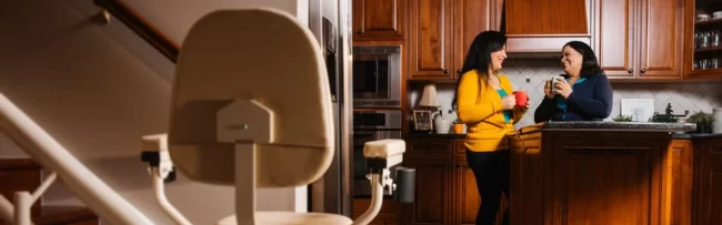 Two women chat in their kitchen with a stairlift in the foreground.