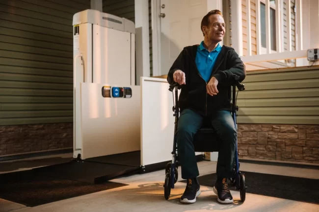 A man sits in his rolling walker next the the vertical platform lift installed at his home.